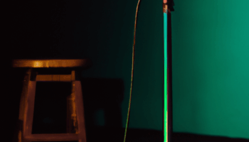 A microphone sites in an empty room with green walls. A brown stool sits in the background.