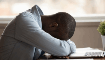 This image shows a person slumped forward over a desk, resting their head on folded arms in front of an open laptop. The person appears emotionally or physically exhausted—possibly overwhelmed, stressed, or burned out. The setting suggests a home or office workspace, with a small potted plant nearby and soft, natural light coming through the window in the background.
