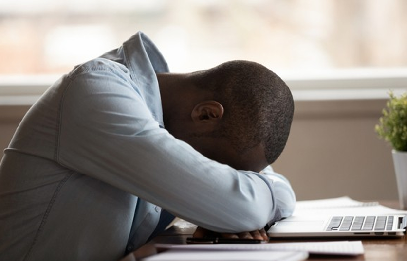 This image shows a person slumped forward over a desk, resting their head on folded arms in front of an open laptop. The person appears emotionally or physically exhausted—possibly overwhelmed, stressed, or burned out. The setting suggests a home or office workspace, with a small potted plant nearby and soft, natural light coming through the window in the background.
