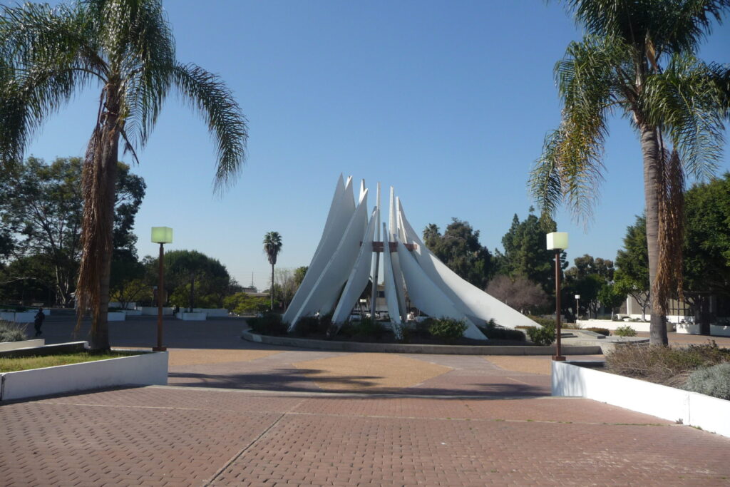 This photo shows a modern sculptural monument surrounded by palm trees under a clear blue sky. The structure features several tall, white, curved panels that rise upward in a circular formation, resembling sails or wings, converging around a central metallic element. The setting is open and landscaped, with a paved circular area and some benches and vegetation around it. The clean lines and symmetry of the monument give it a serene and futuristic feel, set against a backdrop of lush greenery and minimal urban elements.
