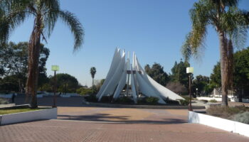 This photo shows a modern sculptural monument surrounded by palm trees under a clear blue sky. The structure features several tall, white, curved panels that rise upward in a circular formation, resembling sails or wings, converging around a central metallic element. The setting is open and landscaped, with a paved circular area and some benches and vegetation around it. The clean lines and symmetry of the monument give it a serene and futuristic feel, set against a backdrop of lush greenery and minimal urban elements.